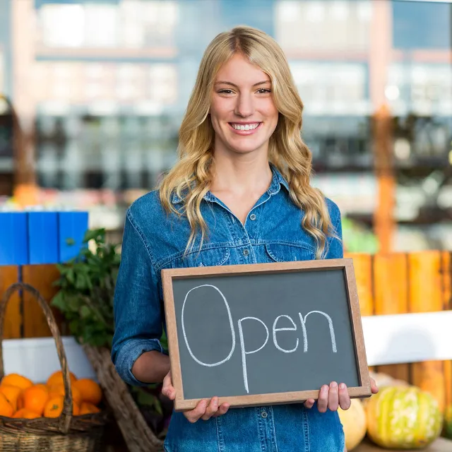 Eine lächelnde Frau steht vor Ihrem Shop mit einem Schild, auf dem "Open" steht